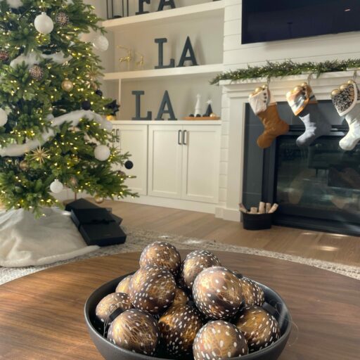 A bowl of bronze animal print Christmas ornaments rest on a table with a Christmas tree, mantel stockings and shelf decorations in the background.
