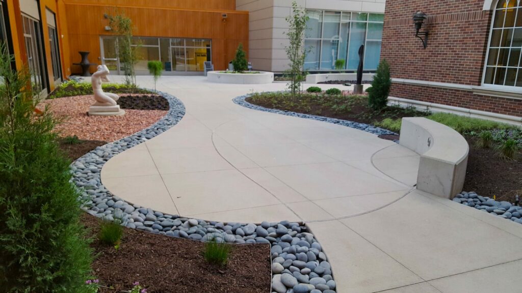Smooth, black river stone borders a flowing, modern concrete hardscape with new plantings and bright sunset orange aggregate color blocks. The client refers to wanting a Japanese influenced garden in her testimonial, and this is a solution that suited the existing courtyard style.