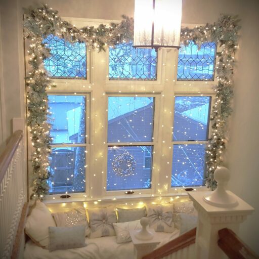A large window is surrounded by upscale lighted Christmas garlands decorated in silver, white and aqua ornamentation. A fairy light curtain is suspended over the entire window. A window seat is covered in a soft, white blanket with nine, elegant white and silver Christmas-themed throw pillows.