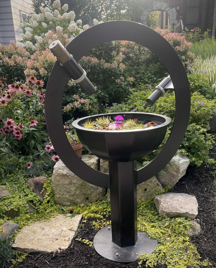 A large, brown steel kaleidoscope sculpture sits in a garden filled with flowering plants. Two scopes point toward a bowl of floating flowers.