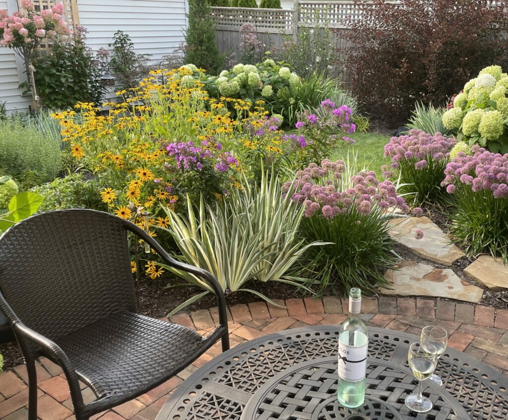White flowering Annabelle Hydrangeas and a pink flowering hydrangea tree are mixed with other flowering perennials in a small backyard garden. A bottle of wine and two wine glasses sit on the iron table in the foreground. 