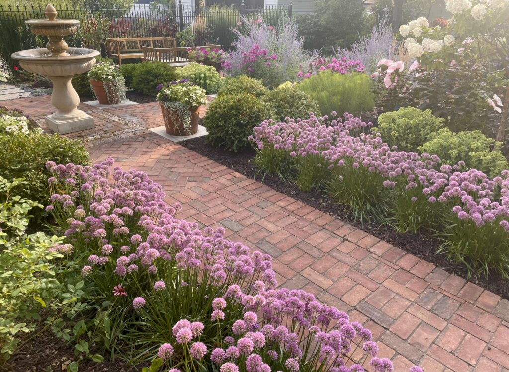 In a testimonial, a client describes that her landscape is unique, custom-made and gives her joy. A basket-weave patterned brick walkway is surrounded by an explosion of blooming plantings. A cast stone, two-tiered fountain sits at the center of a quad of boxwoods surrounded by stone inlay detail in the walkway. A custom-designed iron fence with fleur de lis finials lines the property behind a seating area.
