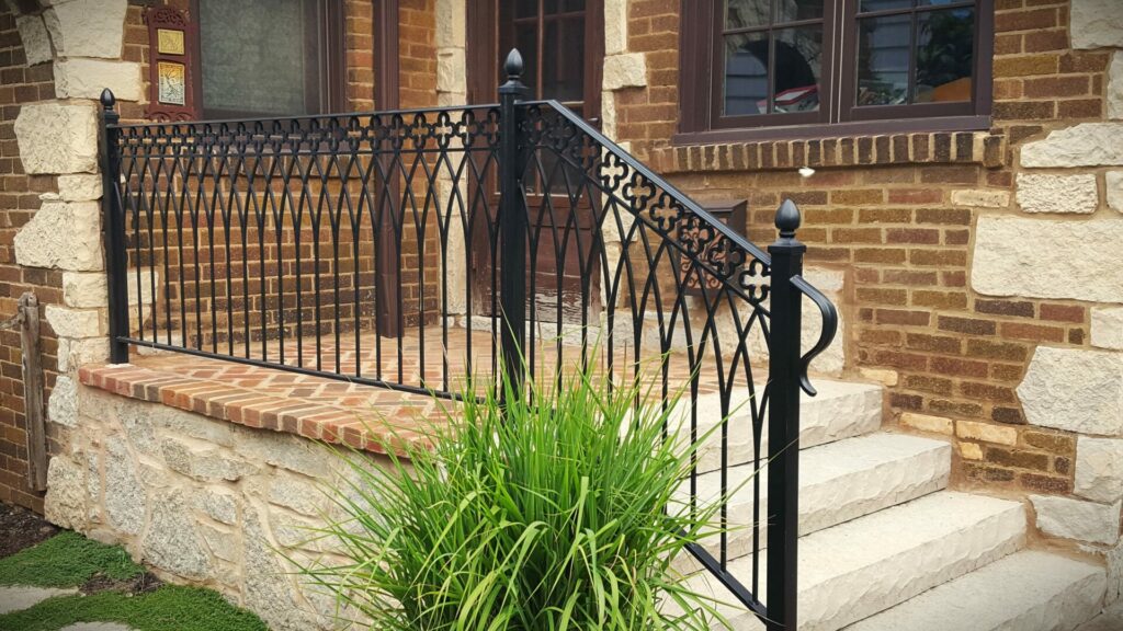 I client describes this custom-made wrought iron railing of gothic arches and rosettes in her testimonial about Bloom! Landscaping. The black iron railing is mounted along four stone steps and a brick stoop set on an irregular stone wall.