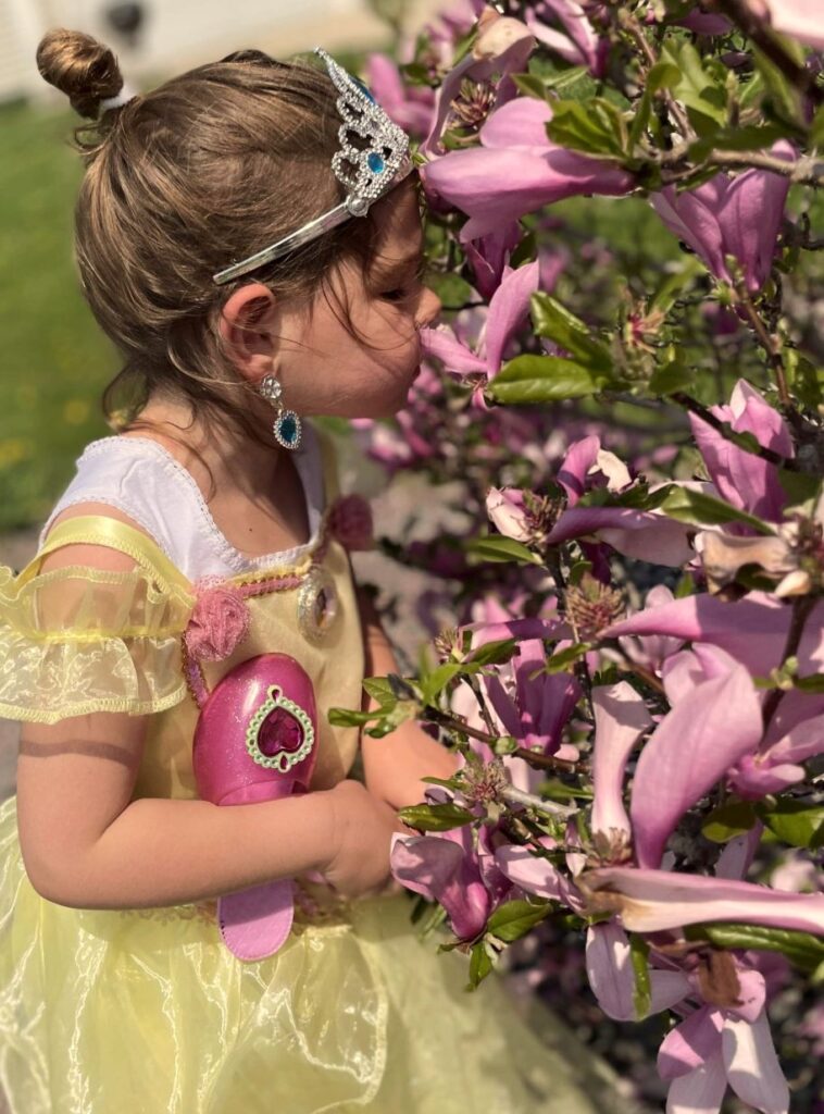 A little girl with her brown hair in a little bun. She wears a yellow princess dress accessorized with a diamond tiara and huge dangling heart diamond earrings. A pink princess slipper with a large heart jewel is tucked under her arm as she passionately smells a pink magnolia flower with her eyes closed. She represents the dream landscapes Bloom! brings to life with well-thought landscaping services near Milwaukee and Waukesha.