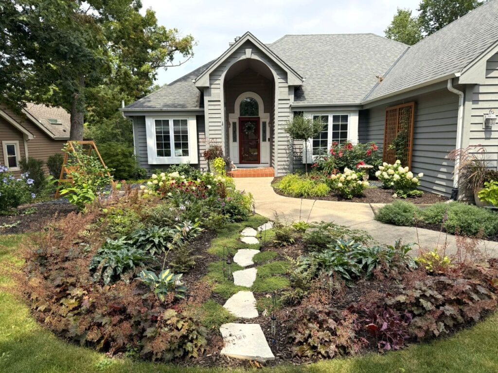 A charming pathway through a shade garden draws the eye to the front entrance of a contemporary gray house surrounded by gardens. The client's testimonial describes the inviting look of her landscape and why she recommends Bloom!